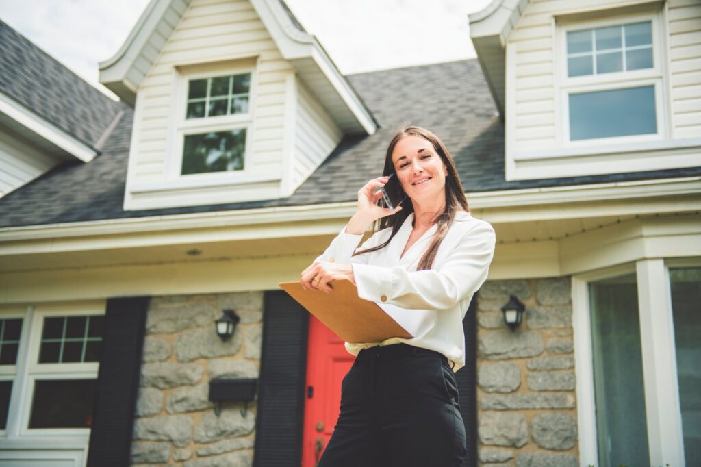 Professional real estate agent talking on the phone while standing in front of a beautiful house.